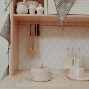 May include: A wooden toy kitchen playset with a white countertop, a wooden pot with a lid, and a wooden stovetop. The kitchen has a shelf with wooden utensils and a wooden cake. The kitchen is decorated with a green and white patterned fabric banner.