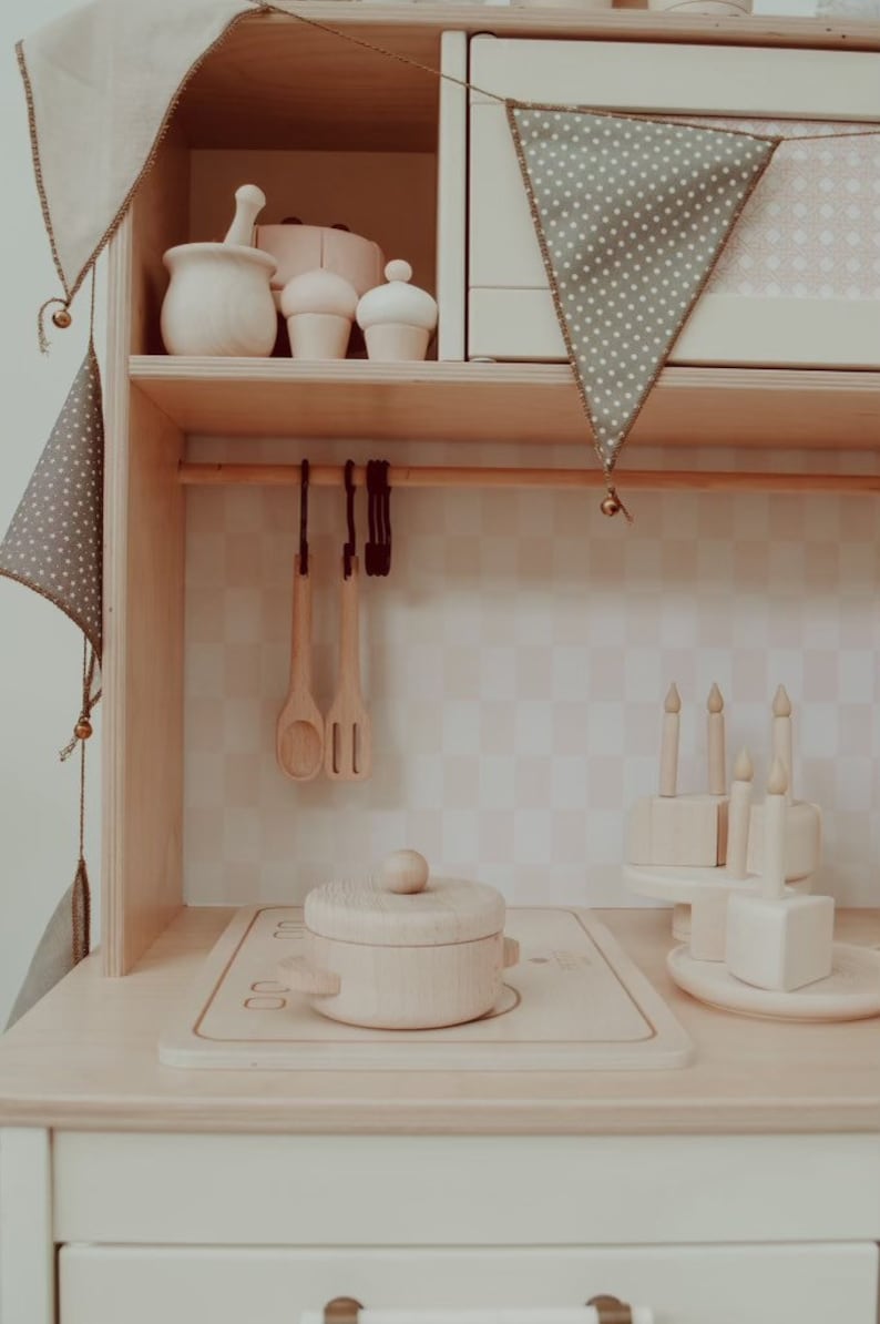 May include: A wooden toy kitchen playset with a white countertop, a wooden pot with a lid, and a wooden stovetop. The kitchen has a shelf with wooden utensils and a wooden cake. The kitchen is decorated with a green and white patterned fabric banner.