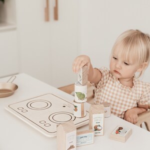 Puede incluir: Un ni&ntilde;o juega con una estufa de juguete de madera y bloques de comida. La estufa tiene dos quemadores y una superficie blanca. Los bloques de comida son de madera y tienen im&aacute;genes de diferentes alimentos. El ni&ntilde;o est&aacute; sosteniendo un bloque con una imagen de un coco.