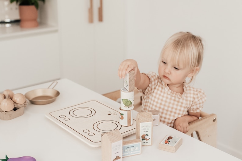 Puede incluir: Un ni&ntilde;o juega con una estufa de juguete de madera y bloques de comida. La estufa tiene dos quemadores y una superficie blanca. Los bloques de comida son de madera y tienen im&aacute;genes de diferentes alimentos. El ni&ntilde;o est&aacute; sosteniendo un bloque con una imagen de un coco.