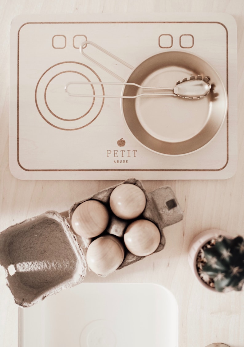 Puede incluir: Una estufa de juguete de madera con dos anillos de quemador y una sart&eacute;n peque&ntilde;a. La estufa tiene un logotipo que dice "Petit Abode". Hay una caja de cuatro huevos de madera al lado de la estufa.