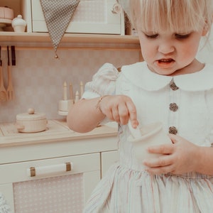 May include: A young girl in a white dress with pink and blue stripes is playing in a toy kitchen. She is holding a white ceramic cup and a wooden spoon. The kitchen is made of wood and has a white oven, a white stovetop, and a wooden shelf with a green and white banner.