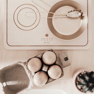 May include: A light-coloured wooden toy stovetop with two burners, a gold-toned metal pan, and a metal serving spoon. The stovetop features the text PETIT ABODE with an apple icon. Below, a cardboard carton holds four smooth wooden eggs.
