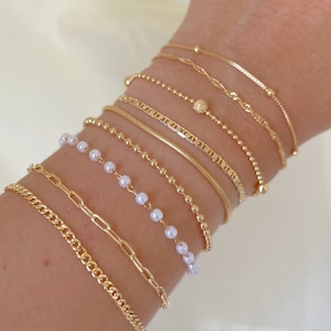 May include: An assortment of gold bracelets, featuring chain, beaded, and pearl designs. The bracelets are shown on a wrist, highlighting their diverse textures and gleam. The jewellery is presented against a soft, neutral backdrop.