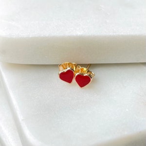 May include: A pair of gold heart-shaped earrings with red enamel. The earrings are small and delicate.