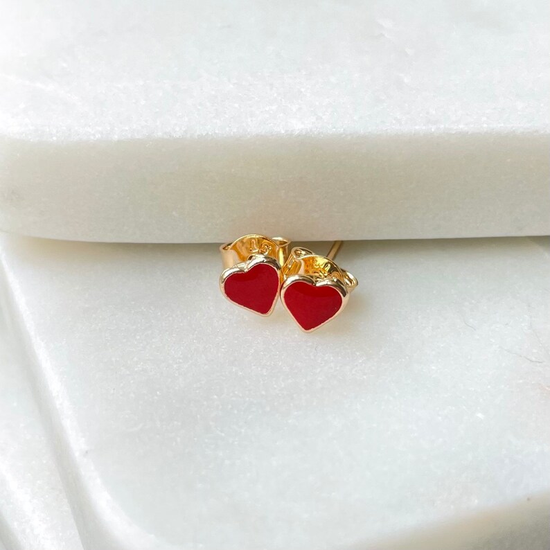 May include: A pair of gold heart-shaped earrings with red enamel. The earrings are small and delicate.