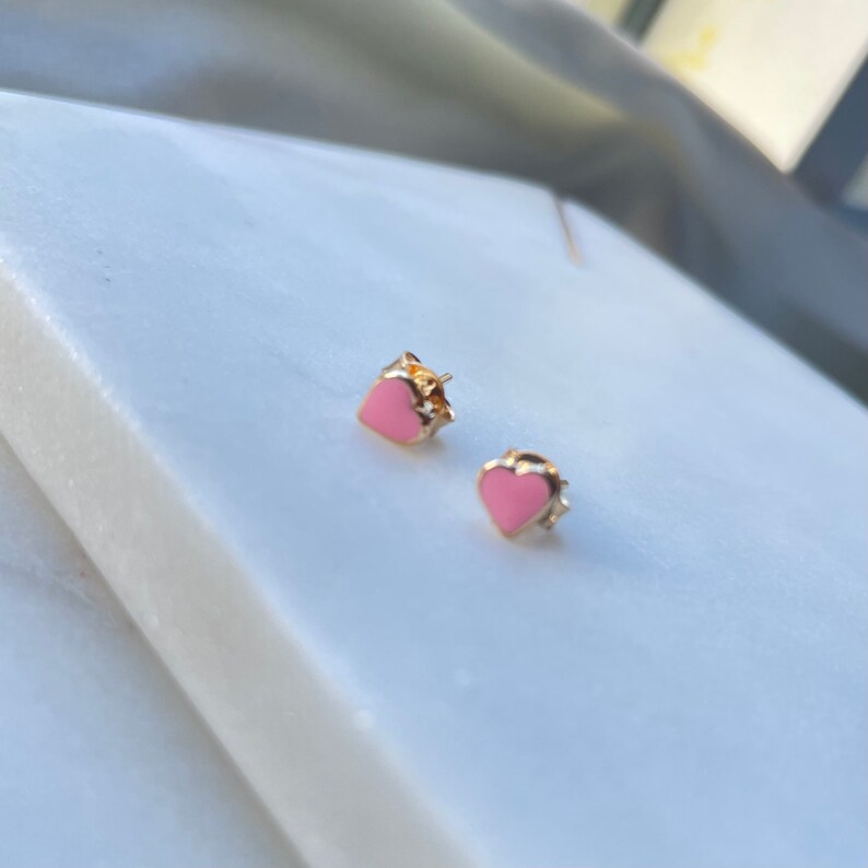 May include: A pair of gold heart-shaped earrings with pink enamel. The earrings are small and delicate.