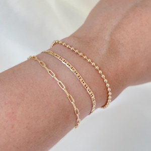 May include: Three gold bracelets stacked on a person's wrist. The top bracelet is a beaded chain, the middle bracelet is a mariner chain, and the bottom bracelet is a chain with oval links.