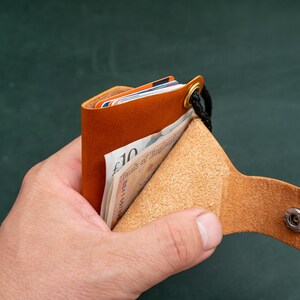 May include: A brown leather wallet with a snap closure and a black strap. The wallet is open and has several cards and a ten pound note inside.
