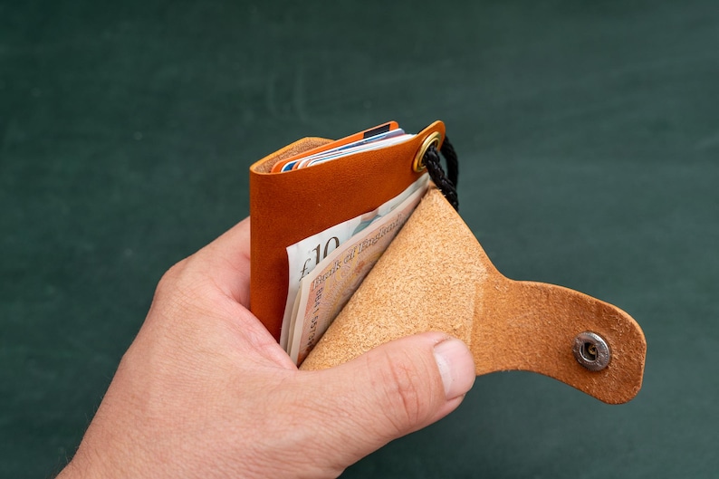 May include: A brown leather wallet with a snap closure and a black strap. The wallet is open and has several cards and a ten pound note inside.