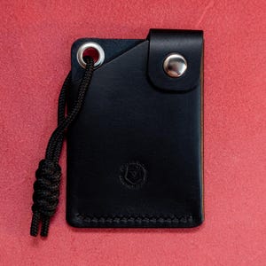 May include: A black leather wallet with a small loop and a silver button. The wallet has a stitched edge and a small logo on the front.