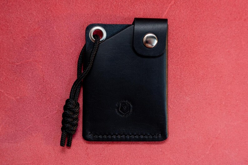 May include: A black leather wallet with a small loop and a silver button. The wallet has a stitched edge and a small logo on the front.