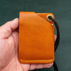May include: A tan leather card holder with a black cord attached. The card holder has a stitched edge and a gold metal eyelet.