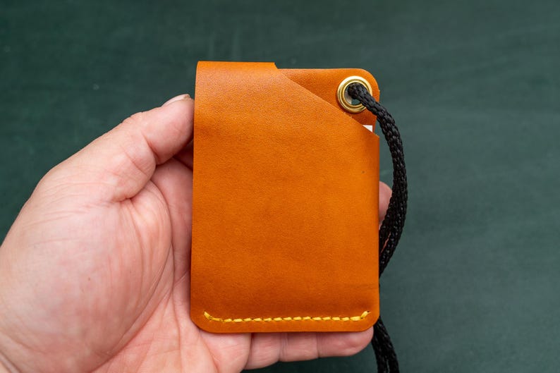 May include: A tan leather card holder with a black cord attached. The card holder has a stitched edge and a gold metal eyelet.