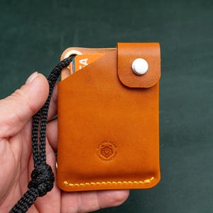 May include: A tan leather card holder with a black cord attached. The card holder has a white button and a small logo on the front.