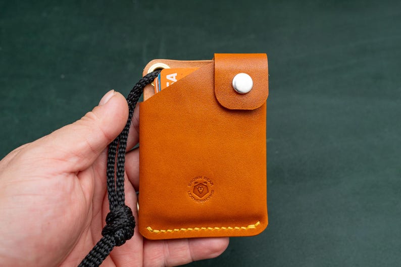 May include: A tan leather card holder with a black cord attached. The card holder has a white button and a small logo on the front.