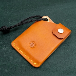 May include: A small, brown leather wallet with a black cord attached. The wallet has a flap with a snap closure and a stitched edge. The wallet has a logo embossed on the front.
