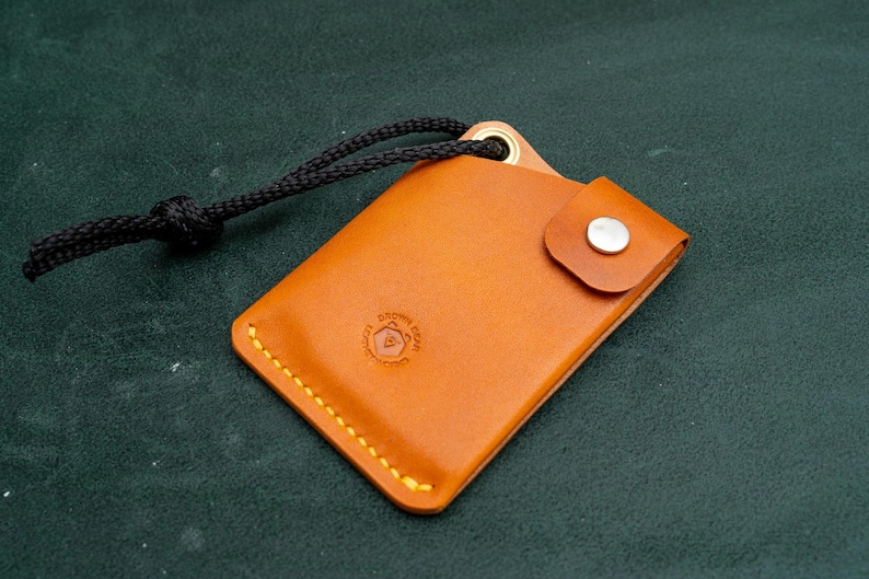 May include: A small, brown leather wallet with a black cord attached. The wallet has a flap with a snap closure and a stitched edge. The wallet has a logo embossed on the front.