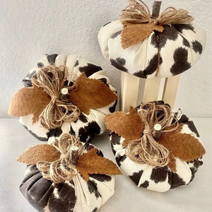 May include: Four fabric pumpkins with a brown and white animal print design. Each pumpkin has a brown felt leaf and a burlap bow.