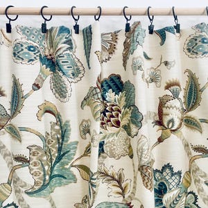 May include: A cream-colored curtain panel featuring a large-scale floral pattern in shades of teal, olive green, and brown. The fabric is suspended from a wooden rod by eight black metal curtain rings with clips, creating soft, vertical folds.