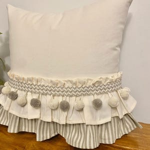 May include: A square off-white throw pillow featuring a horizontal band of textured cream trim adorned with alternating light gray and cream pom-poms. The lower section of the pillow is finished with a ruffled skirt in a light gray and white stripe.