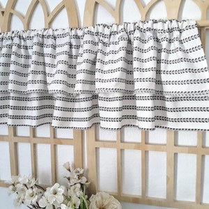 May include: A white valance with a repeating black leaf pattern. The valance is layered and gathered at the top, creating a ruffled effect. It is displayed against a light wooden trellis, with white flowers in the foreground.