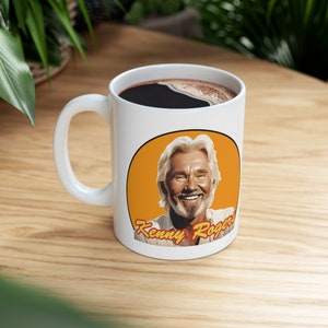 May include: White ceramic coffee mug with a yellow circle featuring a portrait of Kenny Rogers and the text "Kenny Rogers".