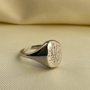 Könnte beinhalten: Ein silberner Siegelring mit einem eingravierten Blumendesign auf der Oberfläche. Der Ring hat eine polierte Oberfläche und ein verstellbares Band. Das Blumendesign zeigt zarte Stiele und Blätter.