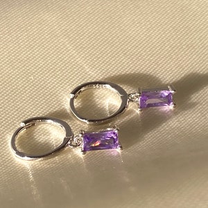 Puede incluir: Un par de pendientes de aro de plata con una piedra preciosa rectangular de color púrpura que cuelga.