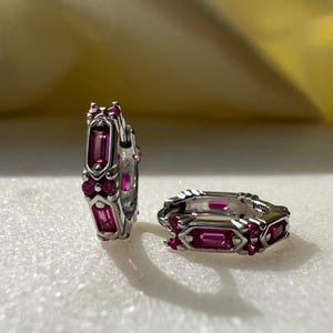 Könnte beinhalten: Ein Paar silberne Creolen mit rosafarbenen Edelsteinen. Die Ohrringe sind klein und zart, und die Edelsteine sind in einer Zargenfassung gefasst.