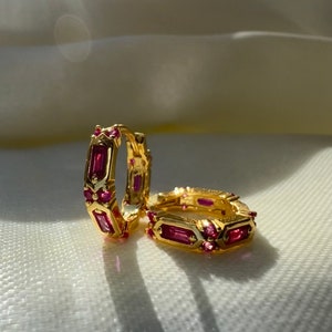 Op de afbeelding: Een paar gouden oorringen met roze rechthoekige edelstenen. De oorringen liggen op een wit oppervlak.