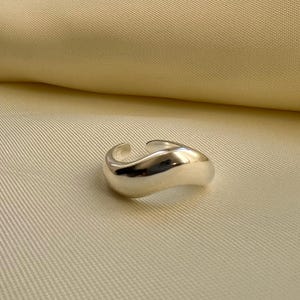 Könnte beinhalten: Ein silberner Ring mit einem glatten, geschwungenen Design. Der Ring ist aus Metall und hat einen schlichten, modernen Stil.