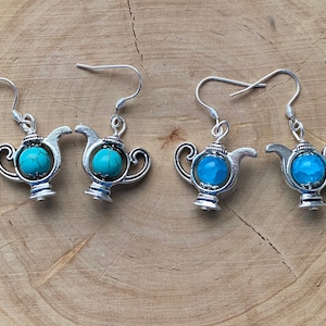 Op de afbeelding: Zilverkleurige theepot oorbellen met turquoisekleurige stenen accenten. De oorbellen hebben een gedetailleerd theepotontwerp met een gebogen tuit, handvat en een turquoise steen in het midden. De oorbellen worden gepresenteerd op een houten oppervlak.