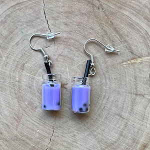Könnte beinhalten: Ein Paar ausgefallene Ohrringe in Form von Miniatur-Bubble-Tea-Getränken. Jeder Ohrring hat eine kleine Glasflasche, die mit lavendelfarbener Flüssigkeit und schwarzen Boba-Perlen gefüllt ist, mit einem schwarzen Strohhalm. Die Ohrringe hängen an silberfarbenen Haken.