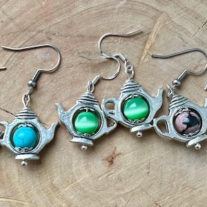 Op de afbeelding: Een verzameling zilverkleurige theepot oorbellen met verschillende stenen accenten. De oorbellen hebben turquoise, groene, roze en andere gekleurde stenen. De oorbellen worden gepresenteerd op een houten oppervlak.