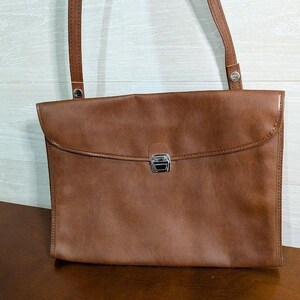 May include: A brown leather shoulder bag with a rectangular shape and a flap closure. The bag has a silver-colored clasp and a long, thin strap. The bag is sitting on a brown surface.