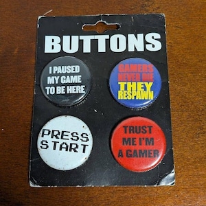 May include: A set of four round buttons on a black card. The buttons have text like "I PAUSED MY GAME TO BE HERE", "GAMERS NEVER DIE THEY RESPAWN", "PRESS START", and "TRUST ME I'M A GAMER".