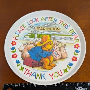 May include: White, round Paddington Bear tray with a colorful illustration. The image features Paddington Bear with luggage and sacks, a train, and the text "PLEASE LOOK AFTER THIS BEAR" and "THANK YOU".