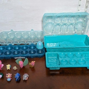 May include: A collection of Hatchimals toys and storage containers. The set includes several small, colorful figures, a blue egg, and various plastic containers in shades of blue. The containers appear to be designed for organizing and displaying the toys.