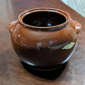 May include: A brown ceramic pot with two side handles. The pot has a rounded shape and a dark interior. The exterior has a glossy finish. The pot is sitting on a dark surface.