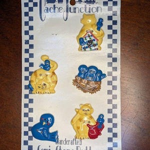 May include: A card of handcrafted semi-stone buttons featuring whimsical yellow cat and blue bird designs. The buttons include a cat holding a birdhouse, a cat with a bird on its head, a bird, a cat with a red bag, and a nest with blue birds. The card reads "Cache Junction".