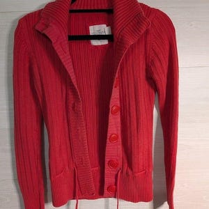 May include: A bright red cardigan sweater with a ribbed texture, featuring a collared neckline and large, matching buttons. The sweater has two front pockets and drawstrings at the bottom. It is hanging on a black hanger.