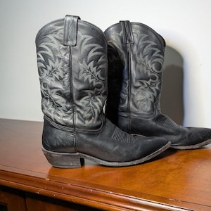 May include: A pair of black leather cowboy boots with intricate light grey floral embroidery. The boots have pointed toes and stacked heels. The leather appears worn, suggesting a vintage or well-loved item.