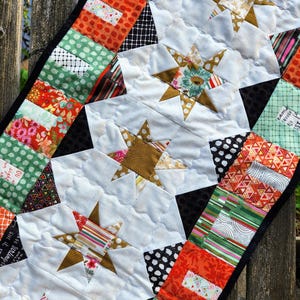 May include: A handmade quilt featuring star patterns and patchwork squares. The quilt showcases a variety of fabrics in shades of white, gold, orange, green, and black. The quilt is bordered with a black fabric.
