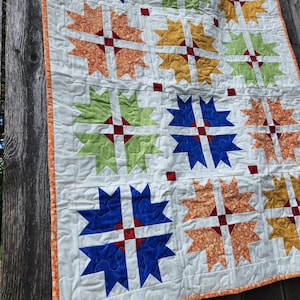 May include: A white quilt with a patchwork design featuring blue, green, orange, and red star-shaped blocks. The quilt is edged with a thin orange trim.