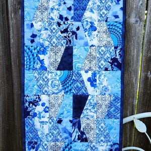 May include: A rectangular, quilted table runner with a patchwork design. The runner features various shades of blue and white fabrics with floral and geometric patterns. A dark blue border frames the design.