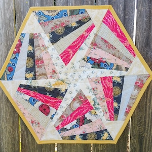May include: A handmade, hexagonal quilted placemat. The design features a starburst pattern with various floral fabric strips in pink, blue, black, and cream. The edges are finished with a gold-coloured binding, creating a decorative and functional item.