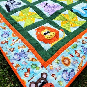 Puede incluir: Una colcha de retazos verde y naranja con fondo azul. La colcha presenta varias caras de animales, incluyendo una cebra, un hipopótamo, un león, una jirafa y un mono. La colcha está rodeada por un borde de tela azul con un patrón de animales y plantas.