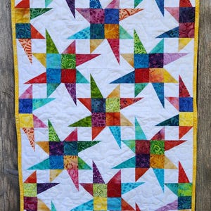May include: A rectangular wall hanging quilt featuring a repeating pattern of multi-colored, pieced star blocks on a white background. The quilt is finished with a bright yellow border and displays intricate, swirling machine quilting stitches.