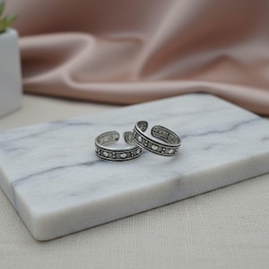May include: Two silver toe rings with a detailed design of small circles and rectangular patterns. The rings are displayed on a white marble surface, with a small succulent plant and soft pink fabric in the background.